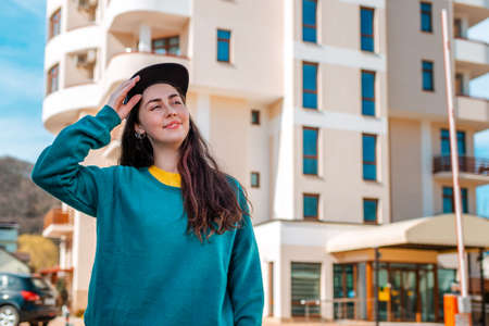 A young Caucasian woman in casual clothes holds her cap with a smile. In the background is a new beautiful multi-storey building.Close up. Concept of buying new housing, mortgages and rentals.の写真素材