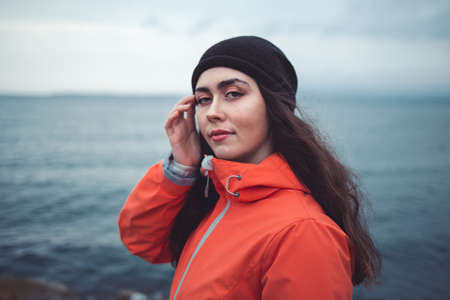 Portrait of a brunette woman with a mysterious look with long hair, wearing a hat and an orange jacket. In the background the sea and the horizon line.の写真素材