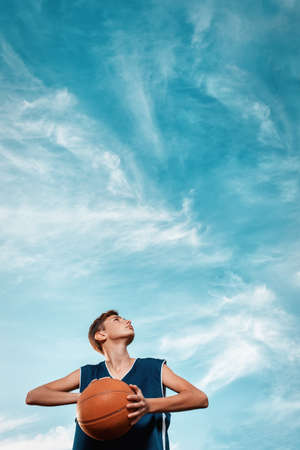 Sports and basketball. A young teen in a black tracksuit playing basketball on the school Playground. Cloudy blue sky on the background. View from the bottom. Vertical. Copy space.の写真素材