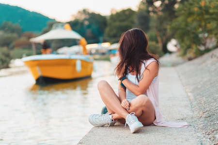 A woman sits cross-legged on a pier by the river, looking at a passing boat. The concept of summer vacation.の写真素材