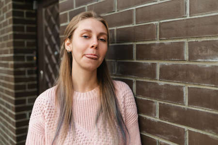 Emotions. Cute young blonde woman shows tongue. In the background, a brick wall.の写真素材