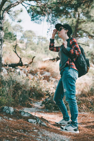 Tourism and outdoor activities. A woman in a cap and glasses, with a backpack on her back, is looking for something in the woods. Vertical orientation.の写真素材