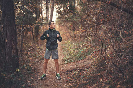Concept of sport and active lifestyle. A young man in a black tracksuit with a backpack on his back, posing in the autumn forest. Dark colors.の写真素材