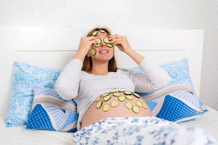 A Caucasian pregnant woman lies on her bed and puts slices of cucumbers on her face and tummy,...