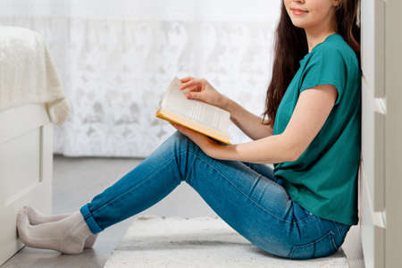 A young Caucasian woman is sitting on the floor and leafing through a book with a smile. Close up. Side view. In the background-the white interior of the room. Concept of education and world book day.の写真素材