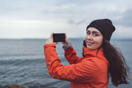 Travel and technology. A smiling woman in a hat and orange jacket takes a picture on her phone. In the background, cloudy sky and sea. Autumn. Copy space.の写真素材