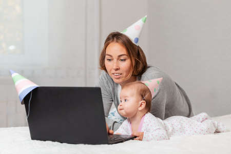 Mother and baby in holiday hats, lying on the bed and using a laptop. Concept of online greetings and video communication.の写真素材