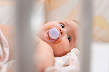 Close up portrait of a baby with a pacifier in his mouth, who is lying in a cradle. View through the bars of the cradle.の写真素材