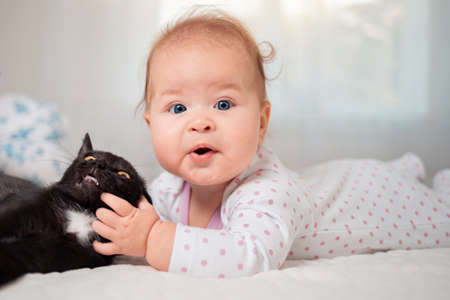 Portrait of a funny baby playing with a cat on the bed. White interior on the background.