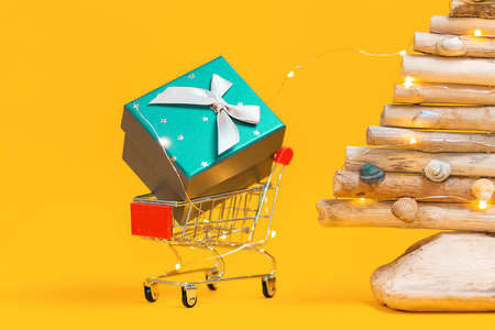 Holiday shopping. A toy shopping cart with a gift, and a craft wooden Christmas tree. Yellow background. Close up. Concept of Christmas sales.