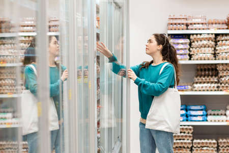 A young Caucasian woman with an eco-bag on her shoulder opens the refrigerator to get food. Shelves with goods in the background. The concept of buying food and shopping.の写真素材