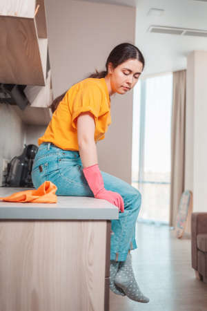 A housewife in casual clothing and rubber gloves sits on the kitchen table, resting after cleaning. Side view.の写真素材