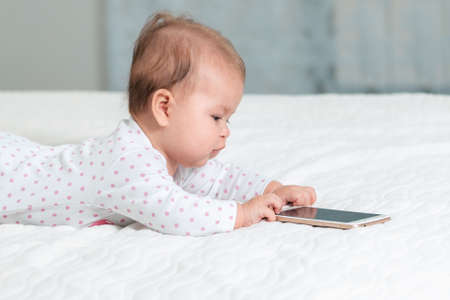 Portrait of baby is lying on the bed and playing with a smartphone. Side view. Copy space. Concept of children's games with modern devices.の写真素材