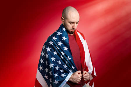 A Caucasian man covered with an American flag stands in a humble and proud pose. Red background. Copy space. Concept of memorial Day, independence day, and other national holidays in the United States.の写真素材