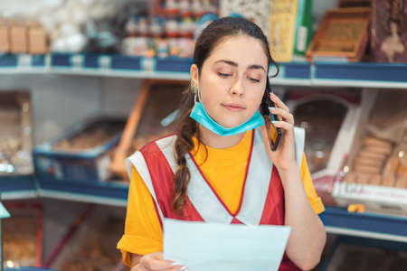 Female salesperson in uniform with a medical mask on her chin, making a phone call making a request for goods. Showcase with products in the background. The concept of doing business during the coronavirus pandemic.の写真素材
