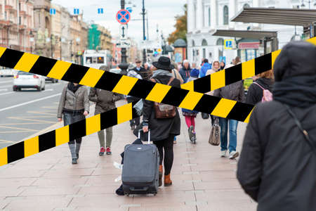 Do not cross.Barrier tape-quarantine, isolation.A girl with a suitcase behind him walking on the sidewalk through a crowd of people.Rear view. The concept of coronavirus and quarantine.の写真素材