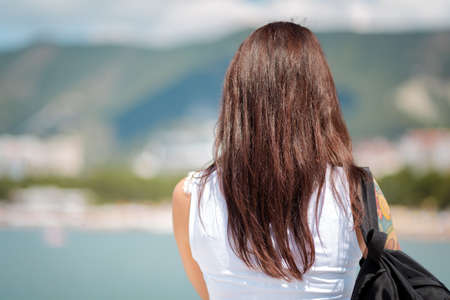 Travel and recreation. A brunette woman looks into the distance. In the background, a view of the mountains and the ocean. Rear view.の写真素材