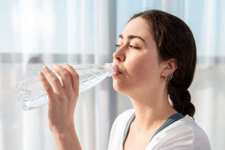 Portrait of a young woman drinking a bottle of water. Large Windows in the background. Healthy lifestyle.の写真素材