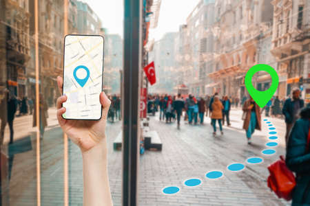 A hand holds a smartphone with an online maps app. In the background, people walking down the street, in a blur. Icon of the route determination. Concept of online maps and travel.の写真素材