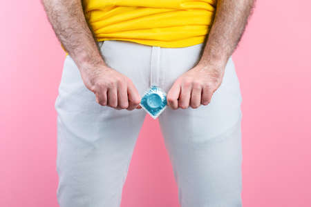 A man in white jeans, legs spread, holds a condom at the level of the genitals. Hands close-up. Pink...