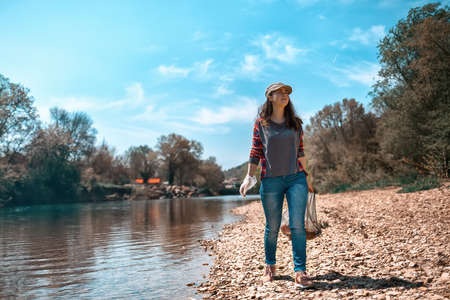 A young woman volunteer collects garbage along the river. The concept of earth day and ecology and environmental conservation. Tint and copy.の写真素材
