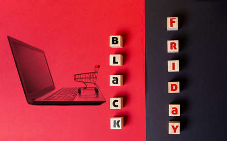 Discounts and sales. On a red and black background are wooden cubes with the inscription black Friday. Flat lay. Image of a laptop with a shopping cart on the red half of the background.の写真素材