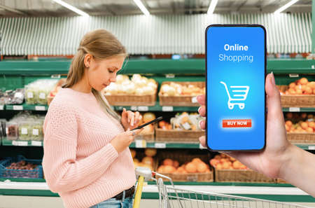 A person's hand holds a mobile phone, with a shopping basket on the screen. In the background, a...