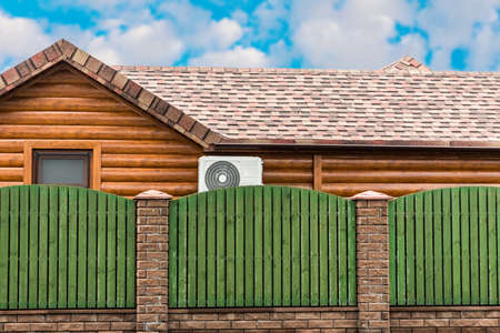 Wooden house with air conditioning, surrounded by a green fence. Blue sky with clouds. General plan.の写真素材