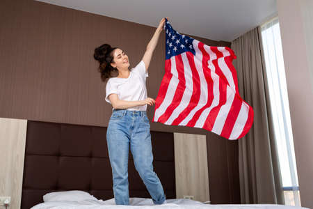 A young happy woman waves an American flag over her bed. Indoor. Concept of the American national holidays and patriotism.の写真素材