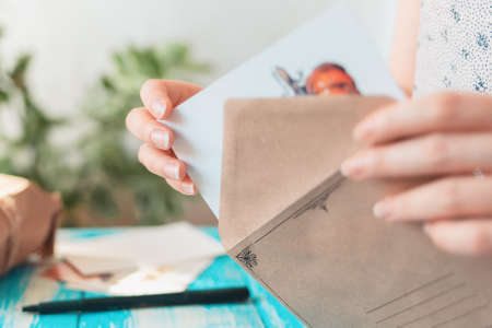 A woman takes a postcard out of a craft envelope. Close-up of envelope. Blue wooden table with a pen in the background. The concept of postcrossing.の写真素材