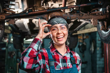 Portrait of young happy repairwoman, in a uniform and glasses poses standing under a car on a lift. Indoors garage.の写真素材