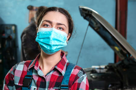 Close up portrait of a young female mechanic in uniform and medical mask. In the background is a car with an open hood.の写真素材