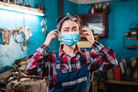 Portrait of a young female mechanic in uniform, wearing a medical mask. In the background are tools and a garage workplace.の写真素材