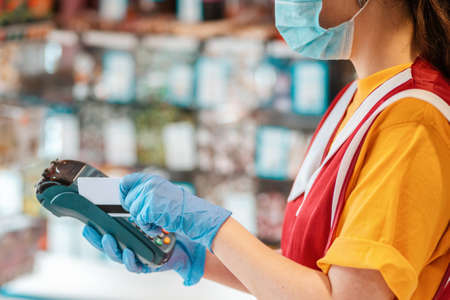 Portrait of an female worker in a uniform with a medical mask on her face and rubber gloves making an online payment. Hands close up. Side view. Concept of online payments by Bank cards.の写真素材