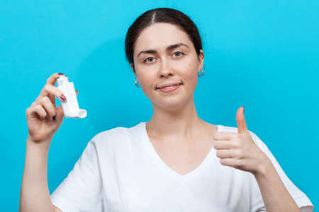 Portrait of a female smiling doctor holding an inhaler in her hand and giving a thumbs up. Blue background.の写真素材