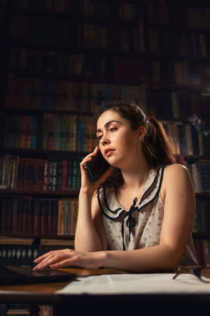 A young woman is working at a computer and talking on the phone. Bookshelves in the background. Evening. Copy space. The concept of self-isolation, remote work and freelancing.の写真素材