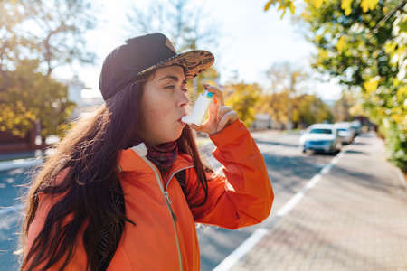 Chronic disease. A teenage girl in casual clothes and cap using an inhaler on the street, having an...