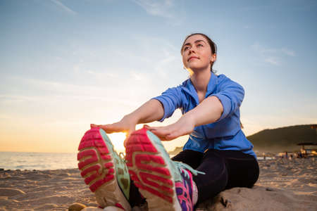 Young woman in sportswear sitting on the sand and performs a stretching of the legs. View from...