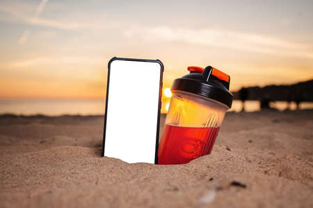 A smartphone and a protein shake shaker are buried in the sand on the beach. Outdoor. Nobody. Mock up. The concept of sports lifestyle and advertising.の写真素材