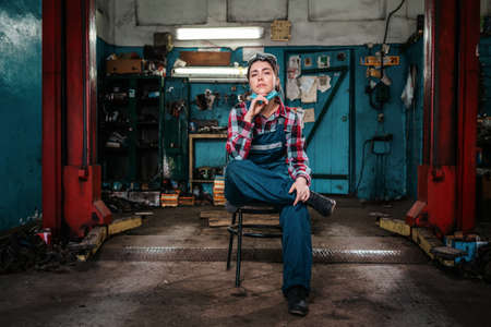 A young female pretty mechanic in a work uniform is sitting on a chair, with a medical mask on her chin. Indoor. Garage.の写真素材