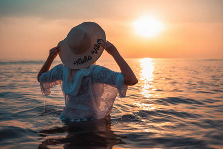 Summer holidays. A woman in a straw hat stands waist-deep in seawater. Sunset. Rear view. Copy space.の写真素材
