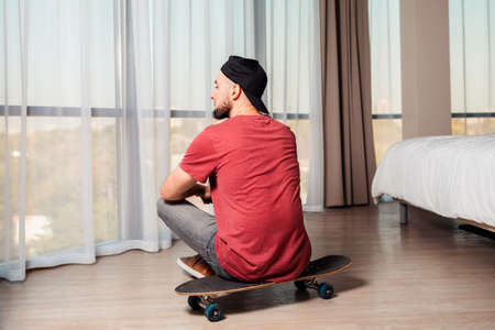 A young man sittingon a longboard, looking out the window. Home interior. Rear view. The concept of quarantine and activity at home.の写真素材
