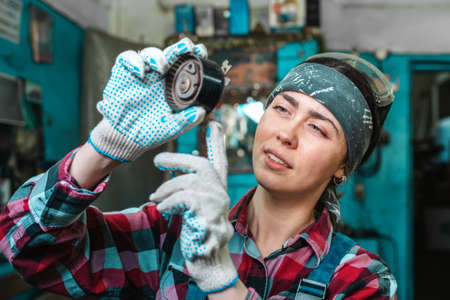 Portrait of a young female mechanic in uniform, wearing gloves and safety glasses on her head, checking a car part. Indoor.の写真素材