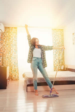 Cleaning concept. Beautiful young brunette woman doing cleaning, dancing with a mop. Sunlight room...