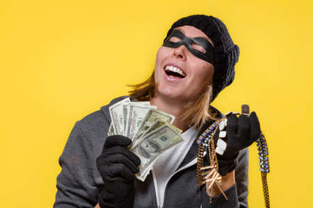 The concept of theft. A woman wearing a hat, mask and gloves holds a wad of dollars and jewelry in both hands. Yellow background and copy space.の写真素材