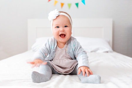 A happy smiling baby girl in a dress and with a headband on her head, sitting on the bed. The...