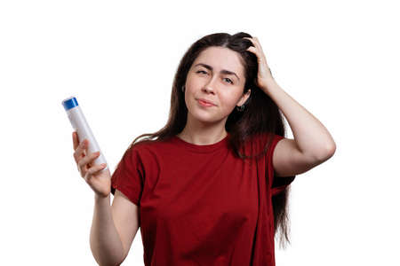 Portrait of a young upset woman who scratches her head and holds a shampoo in her hand. White background. The concept of dandruff and hair care.の写真素材