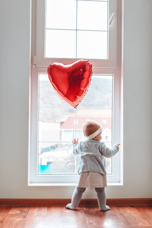 Valentine's day. Little baby girl looks out the window, with a red ball in the form of a heart.