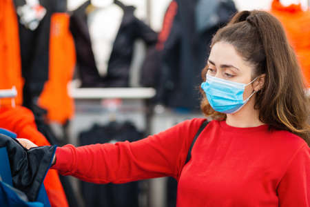 Portrait of a young woman in a medical mask choosing clothes in a store. The concept of shopping during a viral pandemic.の写真素材