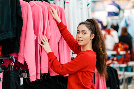 A young beautiful Caucasian smiling woman choosing clothes in a store. Side view. The concept of shopping and consumerism.の写真素材